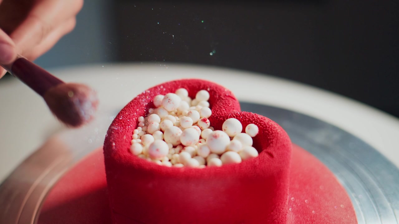 Una mano sparge delicatamente perle bianche su una torta lucida a forma di cuore rosso. La torta è su un supporto girevole, suggerendo una decorazione professionale. Il movimento è lento e preciso, enfatizzando la cura.