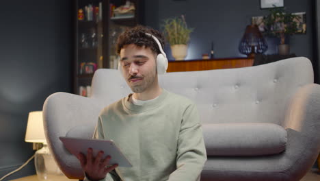 young man watching unexpected movie scene on tablet while drinking soda and sitting with crossed legs on the floor at home 1