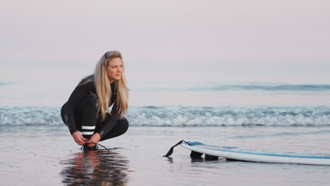 female surfer wearing wetsuit attaching surfboard leash to ankle in waves