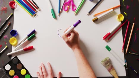 Video-of-hands-drawing-with-purple-pen-on-paper,-with-various-art-materials-arranged-on-table-top