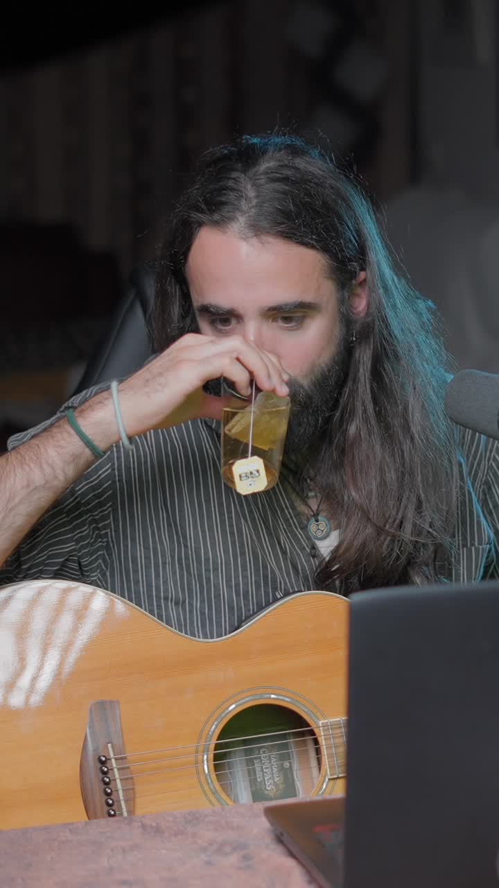 Man with long hair and beard drinking tea while playing acoustic guitar
