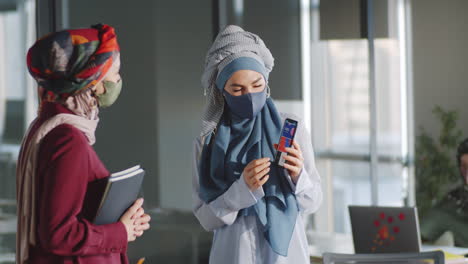 muslim female colleagues in masks using smartphone and speaking