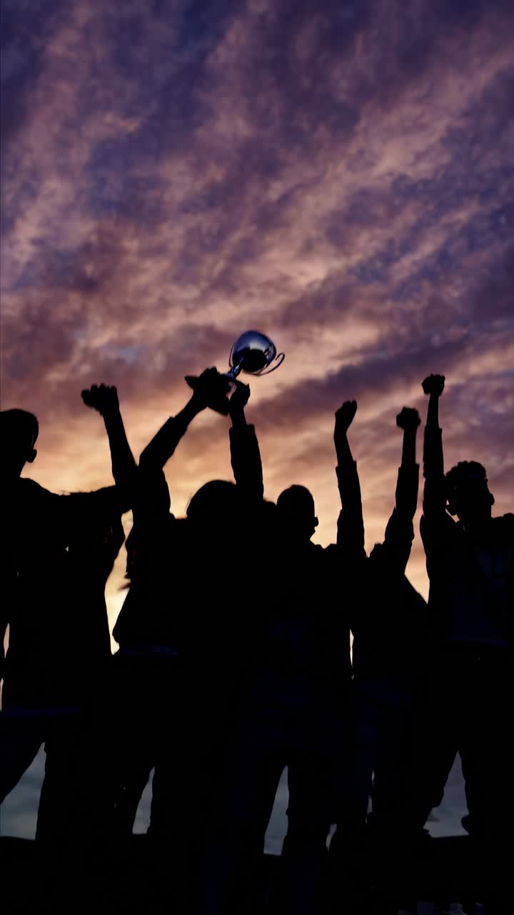Grupo silueteado celebra con un trofeo contra un dramático cielo al atardecer, capturado desde un ángulo bajo.