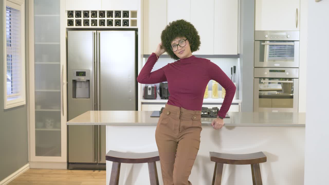 Donna afroamericana appoggiata all'isola della cucina, che si sistema i capelli ricci, incrocia le braccia e ride.