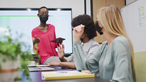 three diverse businesswomen wearing face masks in discussion at office meeting, one holding tablet