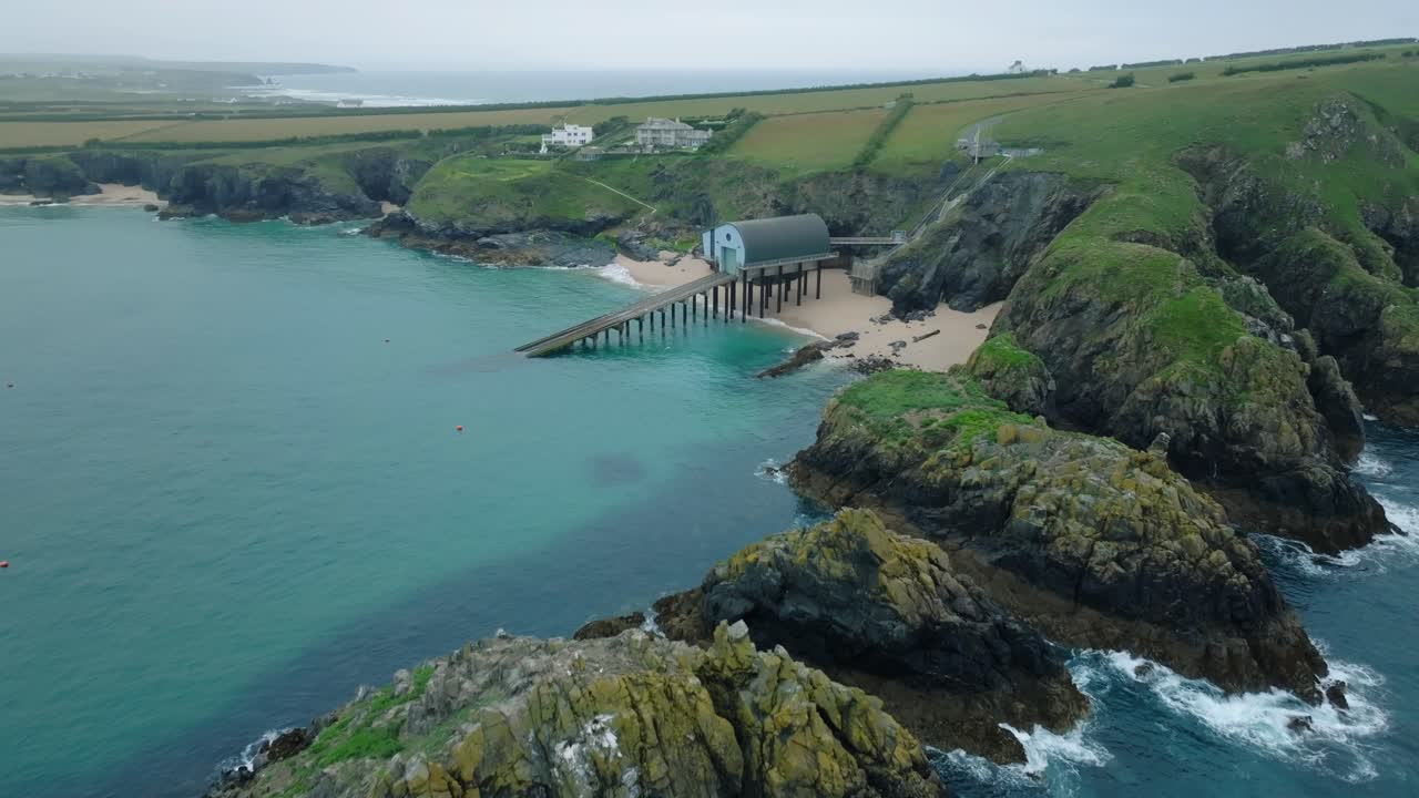 Waves breaking against jagged sea cliffs with camera flight over towards lifeboat station with slipway in clean blue sea. Mother Ivey's Bay, Cornwall, UK.