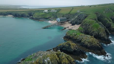 waves breaking against jagged sea cliffs with camera flight over towards lifeboat station with slipway in clean blue sea. mother ivey's bay, cornwall, uk.