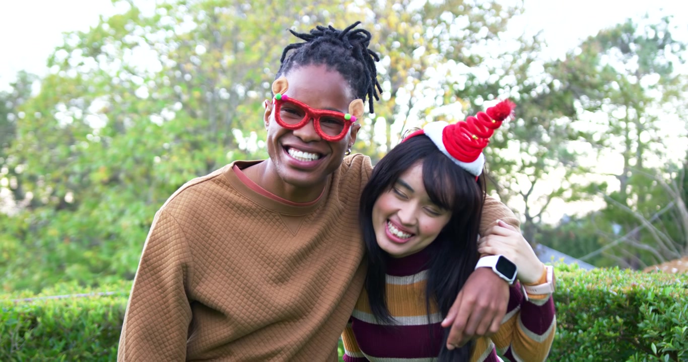 African American man, Asian woman laughing fixing red glasses with headband in park for Christmas
