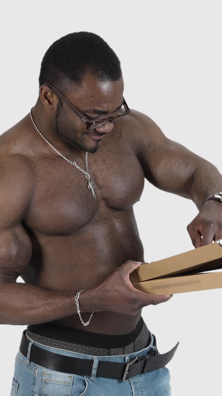 Black skinned sportsman with pizza. Muscled african man in glasses without shirt holding a box and taking out a piece of pizza in studio. Vertical video