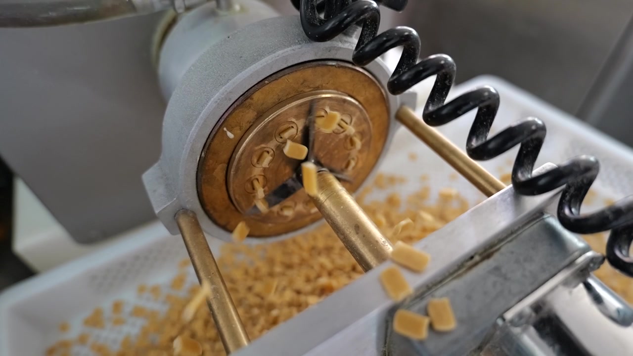 Primo piano dal basso di una macchina industriale per l'estrusione della pasta con una lama da taglio che gira velocemente, che taglia la pasta appena estrusa prima che cada in un vassoio di raccolta
