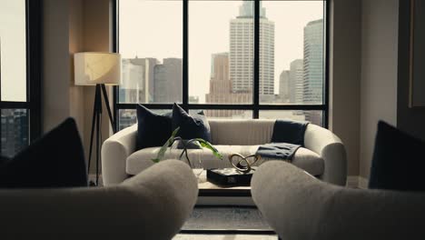 push-in-shot-of-a-living-room-setup-next-to-large-windows-with-a-white-comfy-couch-and-blue-pillows-with-the-city-skyline-in-the-background