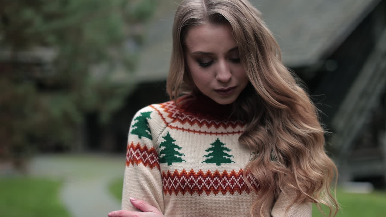 Attractive woman wearing warm sweater posing outdoors. Blonde lady standing outside. Wooden house and nature in blur at the backdrop.