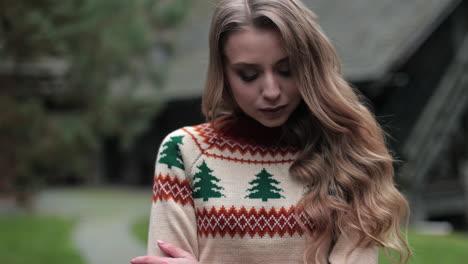 attractive woman wearing warm sweater posing outdoors. blonde lady standing outside. wooden house and nature in blur at the backdrop.