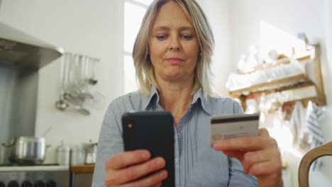 una mujer madura de cabello rubio y ojos azules está ingresando información de tarjeta de crédito usando un teléfono inteligente mientras hace compras en línea en una cocina en casa.