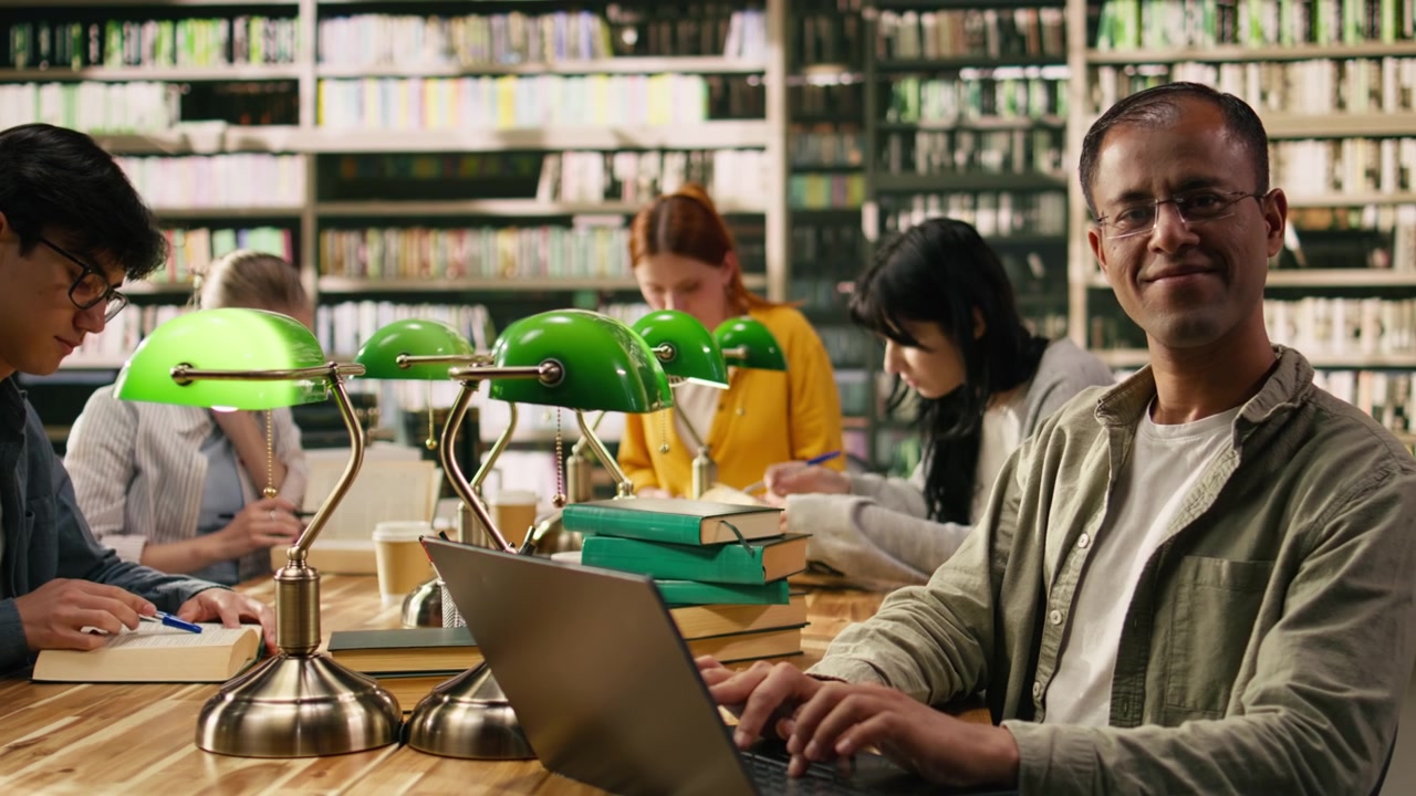 Studente adulto sorridente distoglie lo sguardo dal laptop in una tranquilla area studio della biblioteca.