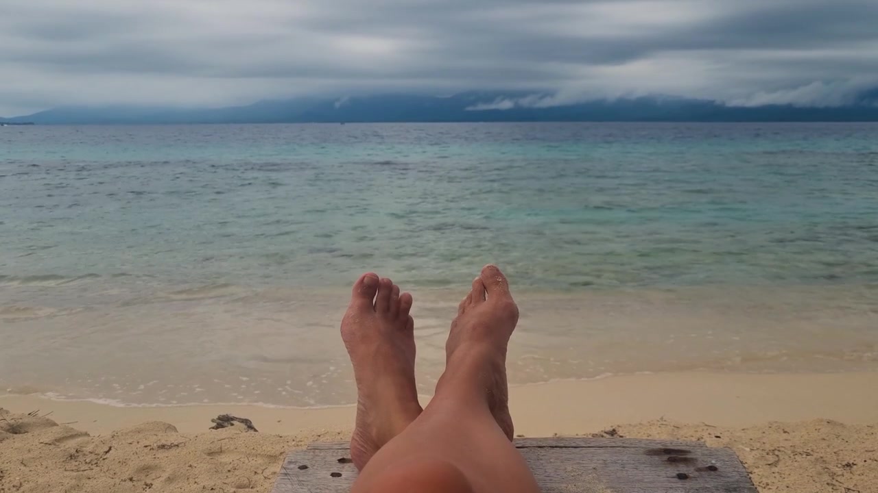 Close-up of woman’s feet relaxing on the beach at Iguana Island, San Blas, Panama, with turquoise waves, empty sand and dark storm clouds gathering over the tropical Caribbean paradise