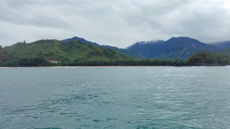 4K-Hawaii-Kauai-Bootfahren-Auf-Dem-Meer,-Das-Von-Rechts-Nach-Links-Neben-Dem-Strand-Schwimmt,-Mit-Bergen-Und-Wolken-In-Der-Ferne-Und-Bootsgischt-Im-Vordergrund
