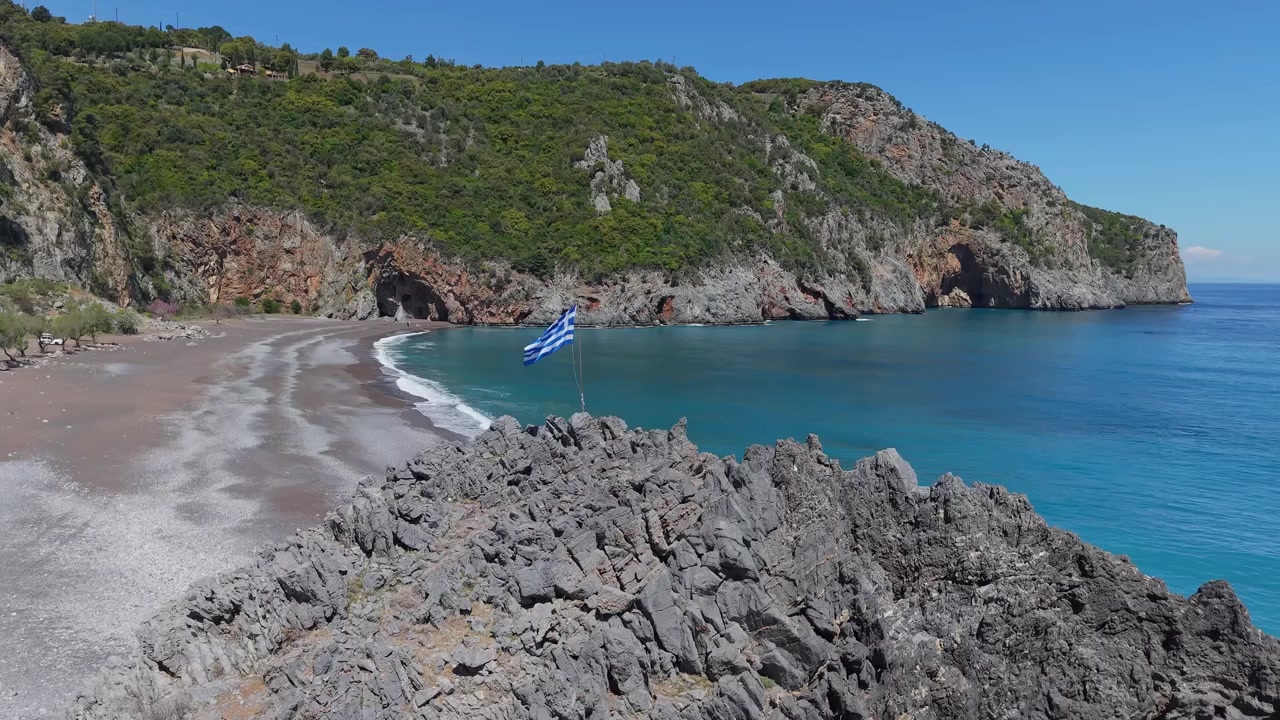 Filmato panoramico di una bandiera greca che sventola sopra la spiaggia di Limnionas, rivelando la costa incontaminata, le acque turchesi e la bellezza aspra della costa dell'isola di Evia