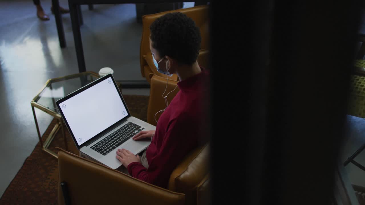 mujer caucásica con máscara facial y auriculares teniendo un video chat en una computadora portátil en una oficina moderna