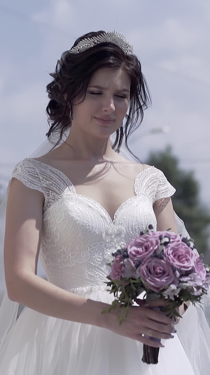 A cámara lenta, una mujer joven y bonita con vestido de novia con escote sostiene un ramo morado frente a edificios de la ciudad