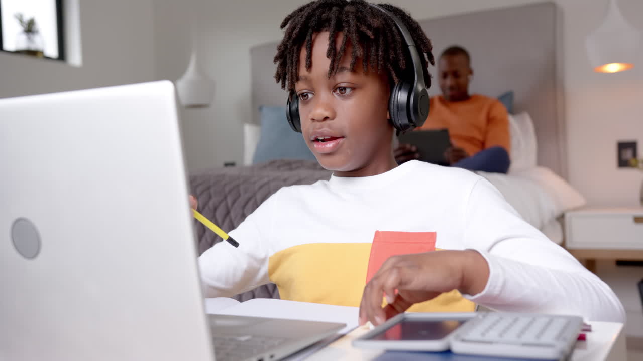 African american boy using laptop for online lesson with father in background at home, slow motion
