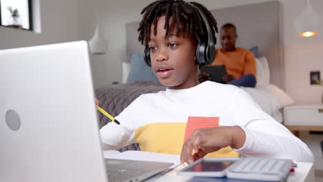 african american boy using laptop for online lesson with father in background at home, slow motion