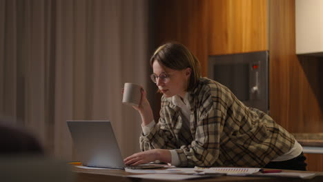 una joven mujer de negocios hermosa trabaja desde casa con una taza de café y mira la pantalla de una computadora portátil de pie en la mesa. ver el correo y beber café. trabajo remoto desde casa