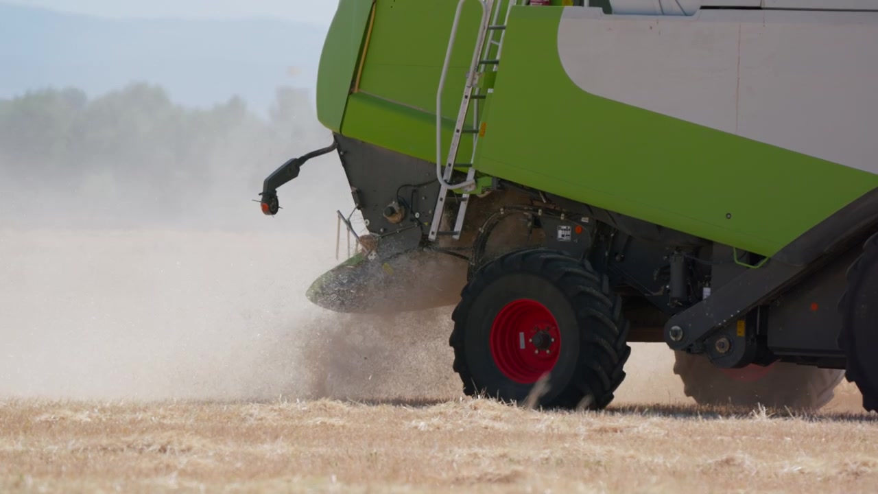 Close up of a green combine harvester discharging dust and straw chaff while harvesting wheat in a dry field, captured in slow motion
