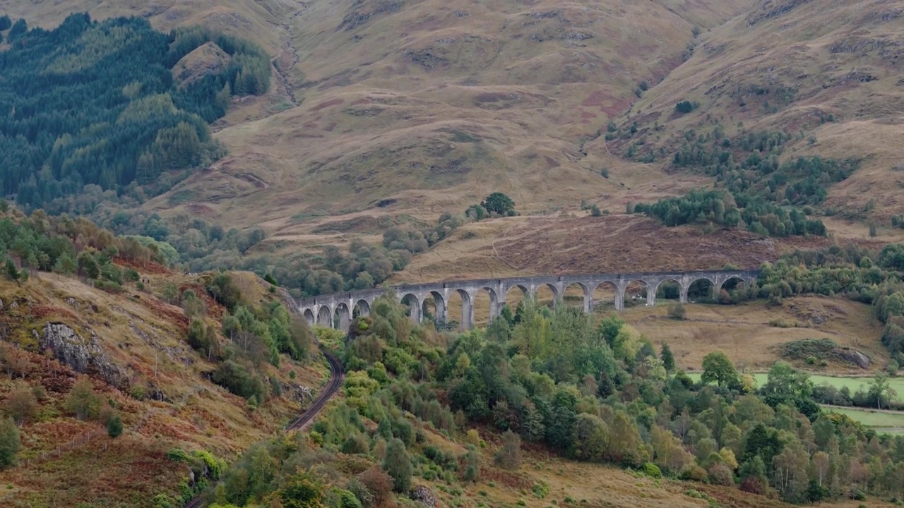 Panorering nedfra og opp av Glenfinnan Viaduct, Skottland, omgitt av bølgende åser og høylandskap, ikoniske togskinner over grønne fjell.