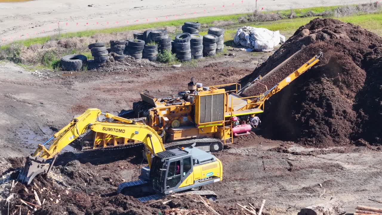 Drone captures an excavator moving soil and mulch in a sunny outdoor setting. Machinery operates efficiently in an industrial landscape