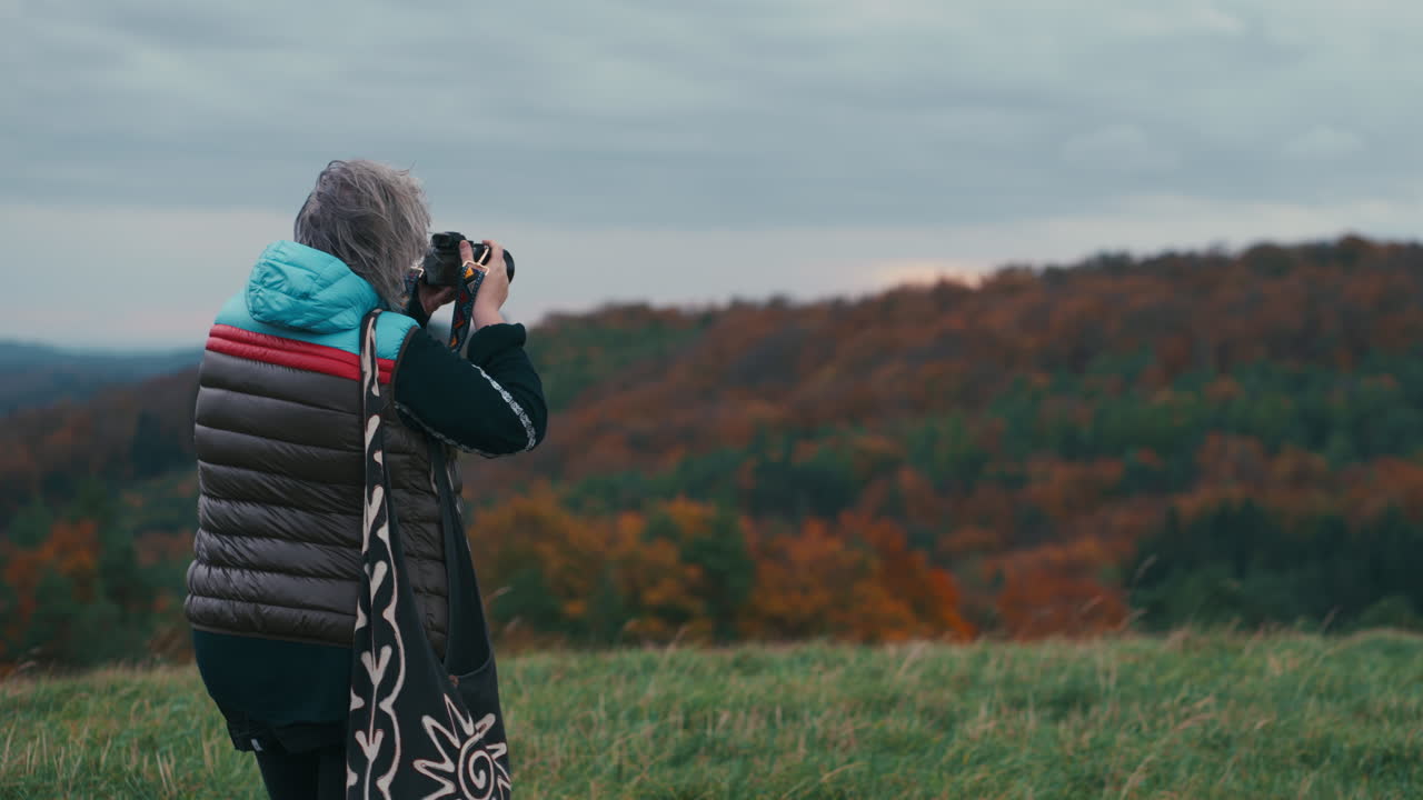 Cinematisk optagelse af en kvindelig fotograf med gråt hår, der tager billeder af et efterårslandskab med sit kamera på en blæsende dag, omgivet af natur, i slowmotion.