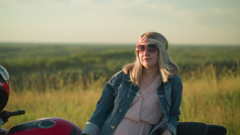 a woman wearing a denim jacket and pink gown smiles while resting on a motorcycle in an open field, she wears sunglasses, giving off a relaxed and carefree vibe