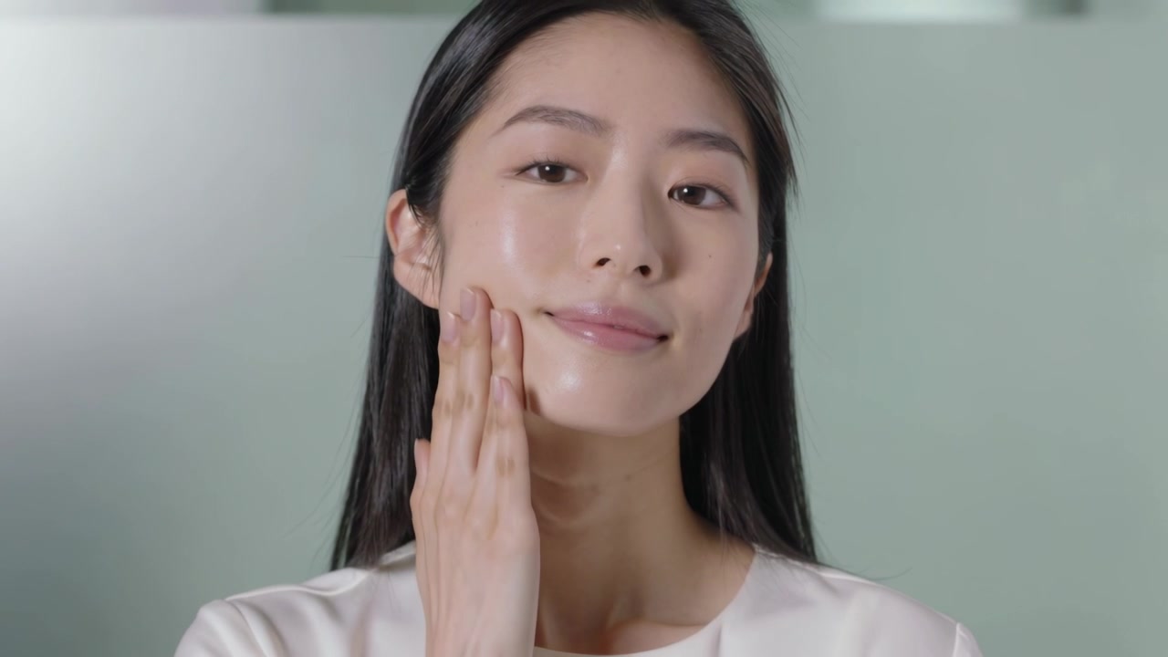 스튜디오에서 흰색 상의를 입은 여자가 세럼을 바른 후 손으로 턱을 위로 쓸어 올리며 볼을 마사지하는 모습.