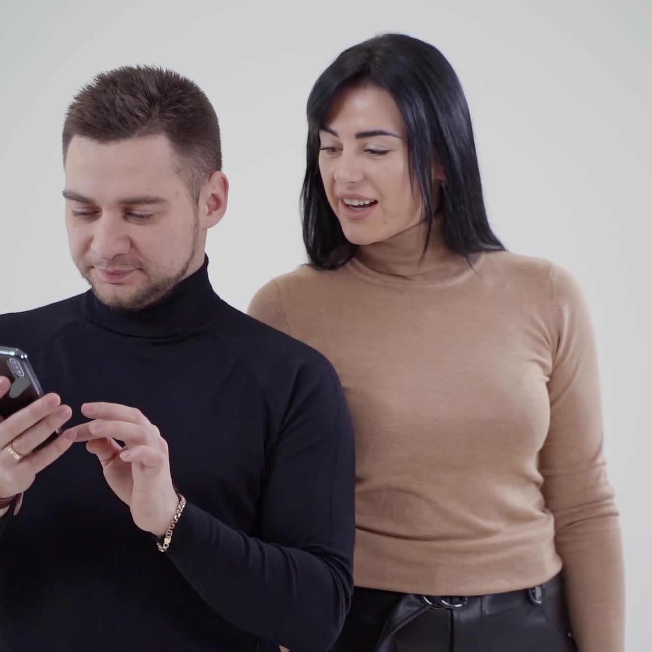 Handsome man looking at the phone in studio. Beautiful young woman standing behind the man. Young couple on the white background.