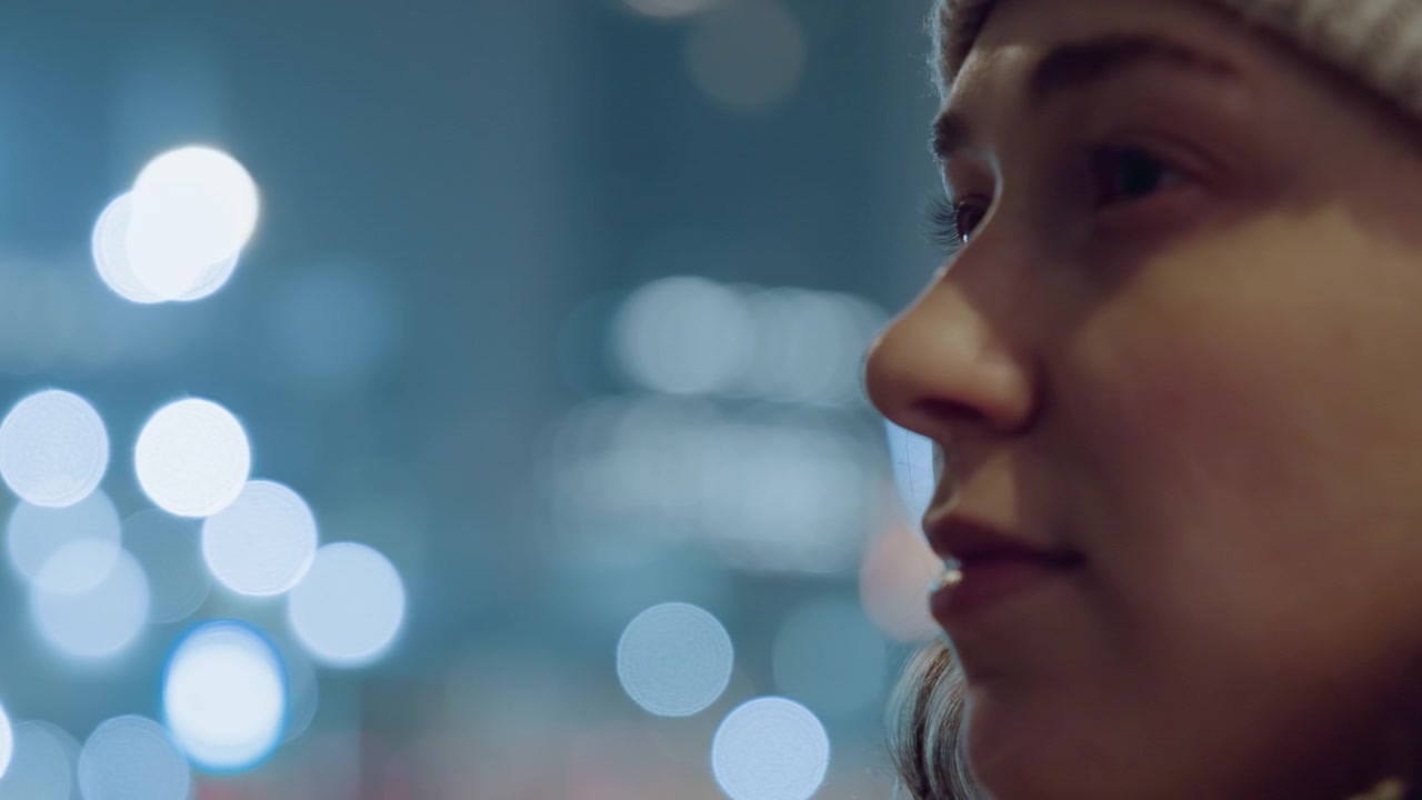 Young caucasian woman adjusting glasses against soft bokeh lights background, close profile framing, beanie and oversized frames, delicate movement, reflective mood, urban evening, cinematic grain