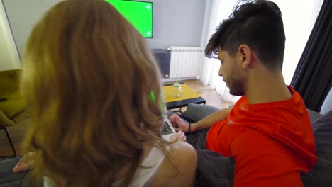 una pareja en la sala de estar con una computadora portátil de pantalla verde y un televisor