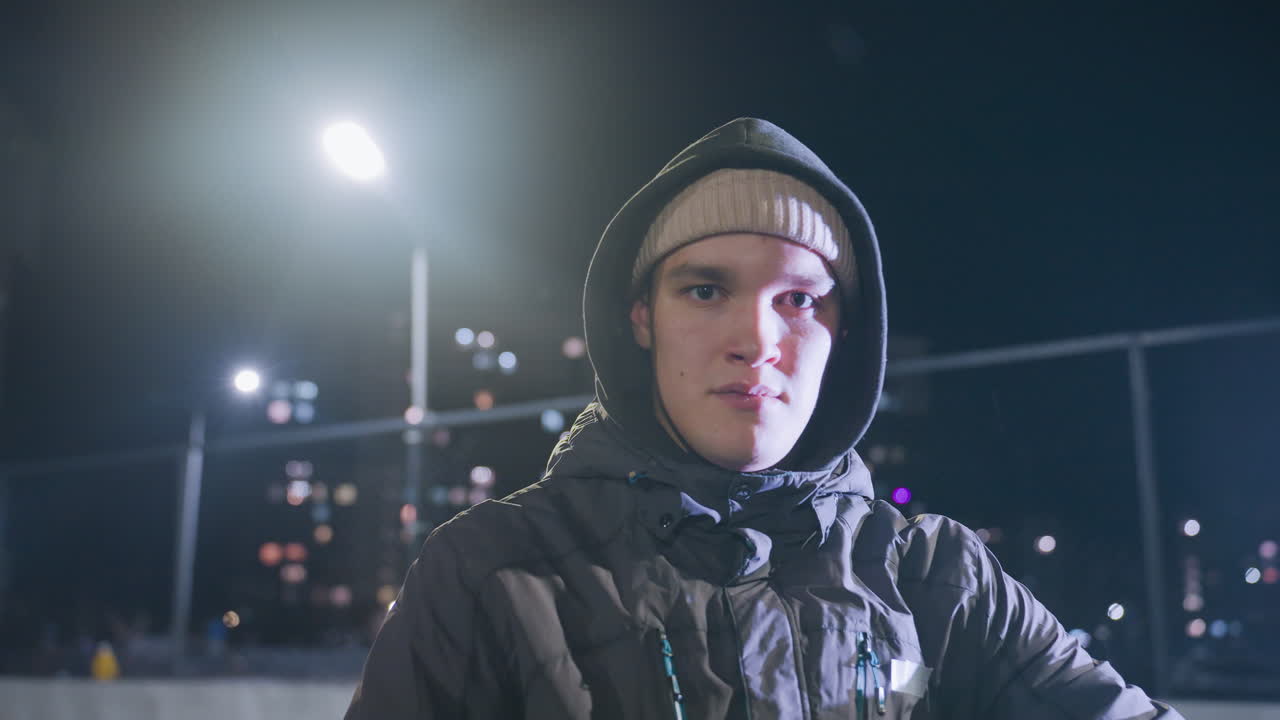 niño joven con capucha sosteniendo la pelota en la mano, mirando pensativo durante la práctica nocturna bajo la luz de la calle brillante con el paisaje urbano en el fondo