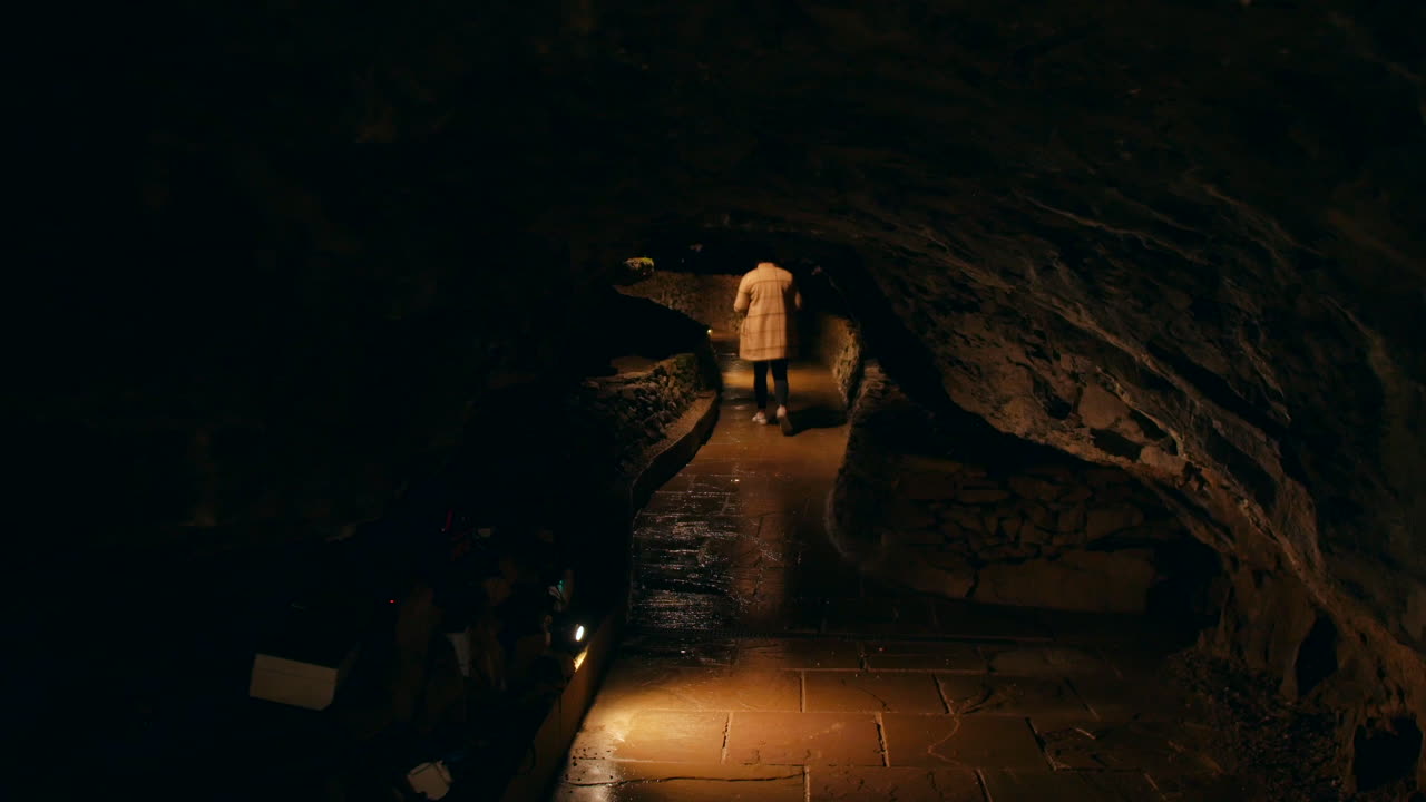 una mujer caminando a través de un túnel subterráneo débilmente iluminado con paredes de piedra ásperas y un techo bajo