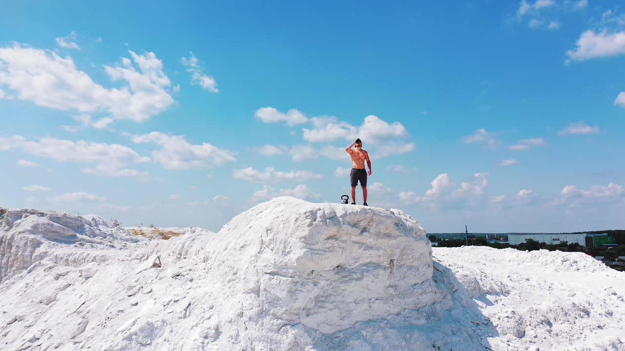 Sporty man drinks water outdoors. Athletic man standing on the top of white hill and drinking after training in summer. Camera moves around.