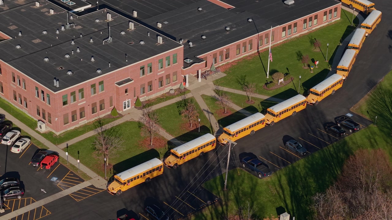 Drone view of yellow school buses lined up outside an American school building in autumn