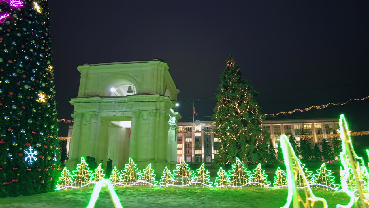 Festive holiday decorations light up the historic square at night. Colorful Christmas lights adorn trees and a beautiful archway, creating a joyful atmosphere for visitors
