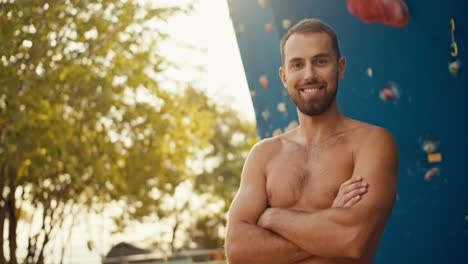 Retrato-De-Un-Escalador-Masculino-Feliz,-Un-Hombre-Moreno-Con-Barba-Incipiente-Cruza-Los-Brazos-Sobre-El-Pecho-Y-Posa-Cerca-De-Un-Muro-De-Escalada-Azul-En-Un-Día-Soleado-De-Verano.