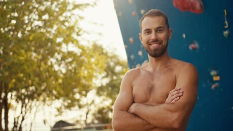 retrato de un feliz escalador de roca masculino, un hombre moreno con paja dobla los brazos en su pecho y posa cerca de una pared de escalada azul en un soleado día de verano