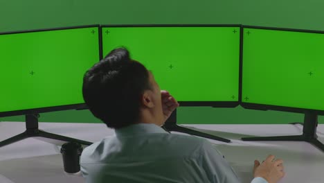 녹색 스크린 스튜디오에서 모의 여러 컴퓨터 모니터와 함께 작업하는 동안 피곤한 아시아 남자의 뒷면