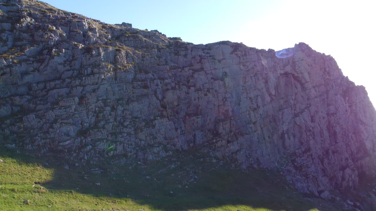 Aerial with Thin Rocky Cliff with Bright Sun Flare as Camera Moves Sideways Revealing Expansive Calm Ocean with Devon Coastline in Far Distance. Travel Nature Drone Clip. Gower Coastline, Wales
