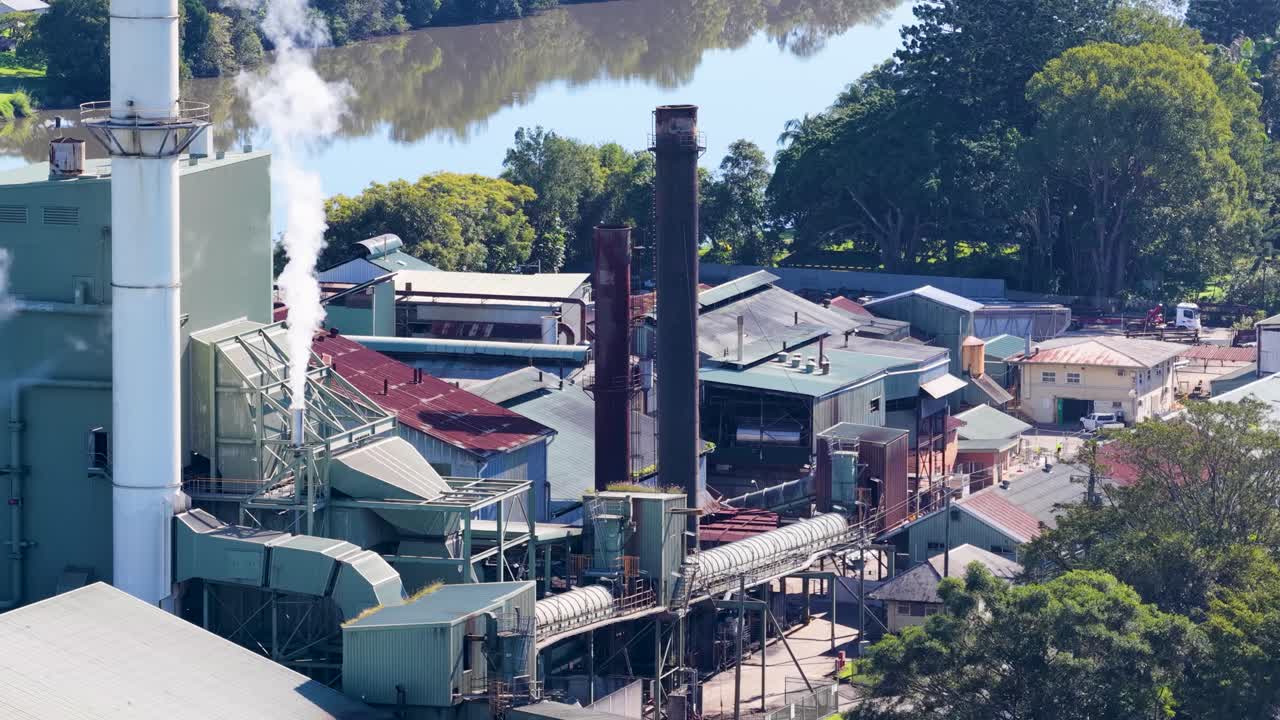 A factory releases smoke into the air, surrounded by trees and buildings, under bright daylight