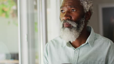 portrait of african american senior man with beard drinking coffee looking out of window and smiling
