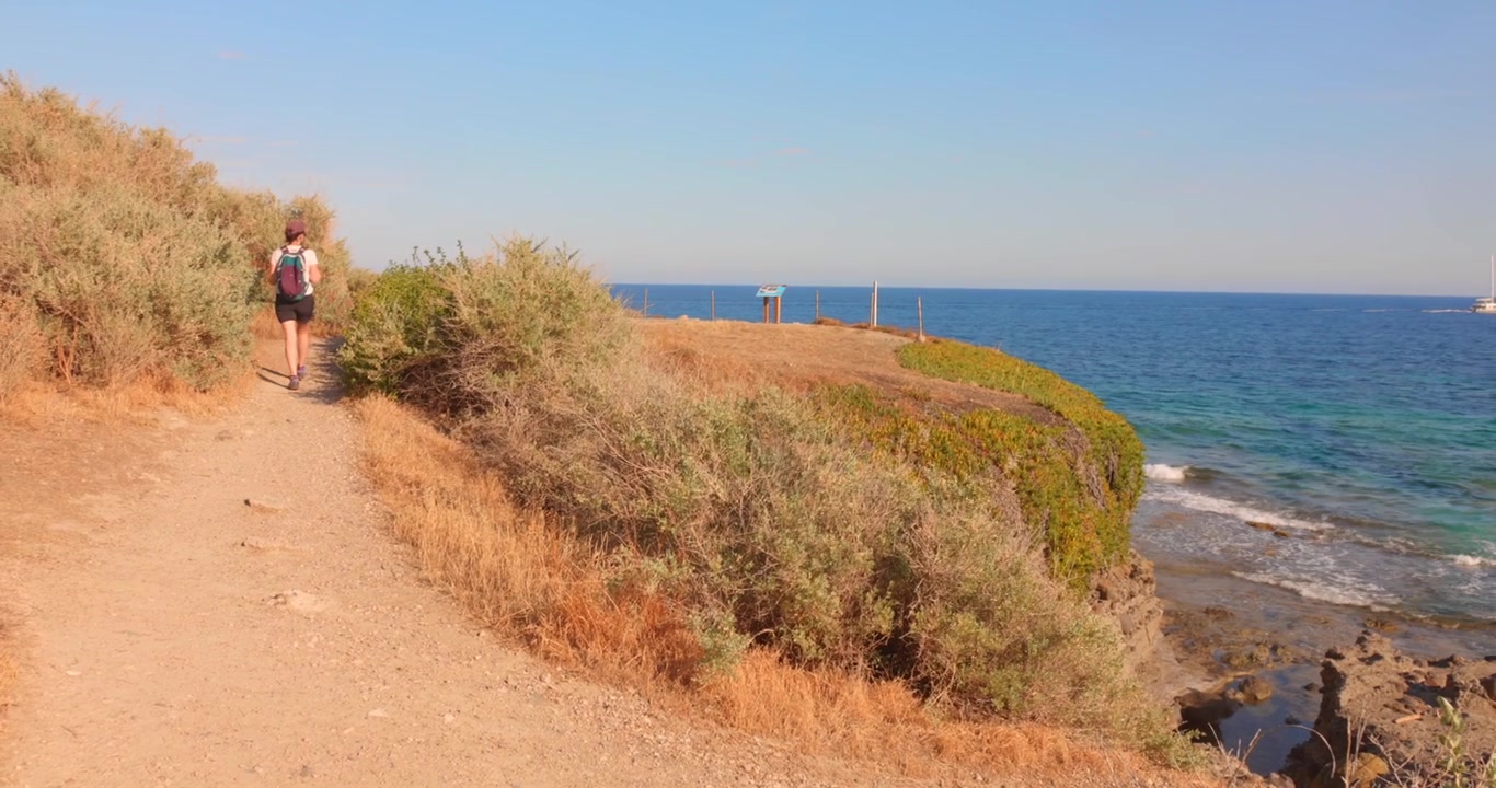 Persona che cammina lungo un sentiero costiero asciutto sopra una costa rocciosa, con il mare blu in vista.