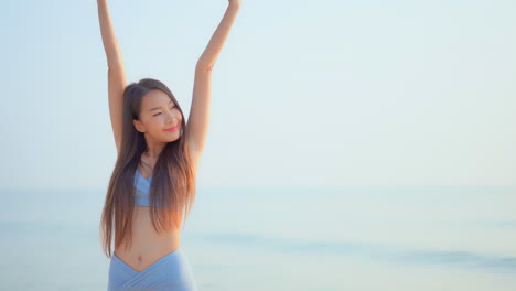 beautiful asian woman model standing in front of the sea looking aside interlock her fingers and raise hands up and spread arms over her head