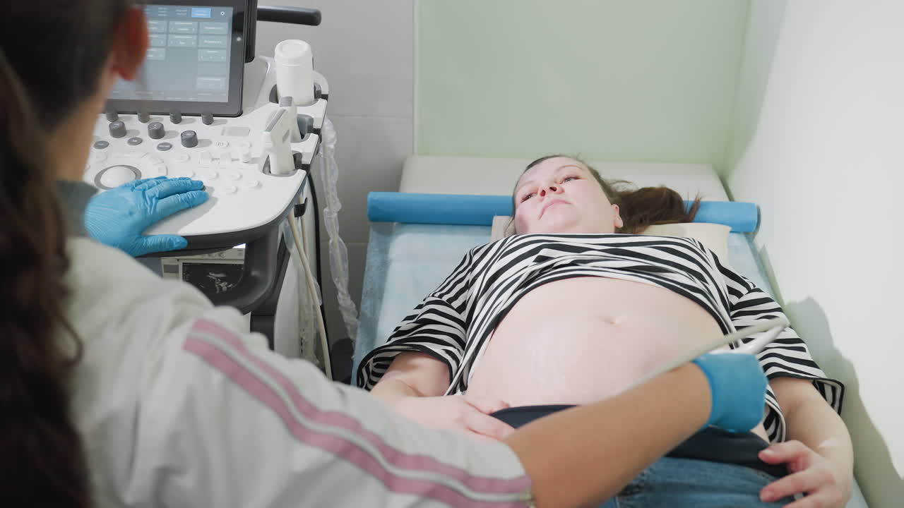 Female medical technologist wearing gloves examines lower abdomen of pregnant woman using ultrasound scanner while simultaneously adjusting control panel during prenatal checkup in clinical room